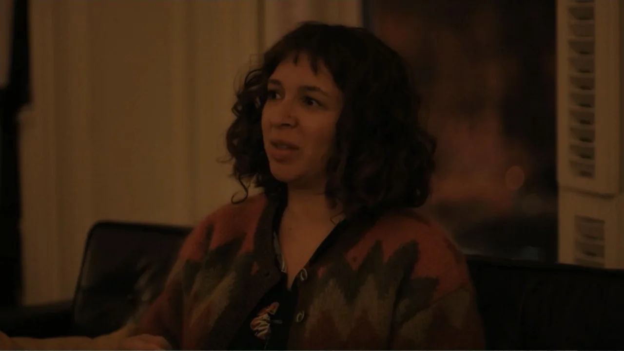 Maggie's Plan (2016) movie still: medium — A dimly lit room with blurred dark walls and a visible window shutter. A woman in a patterned knit…