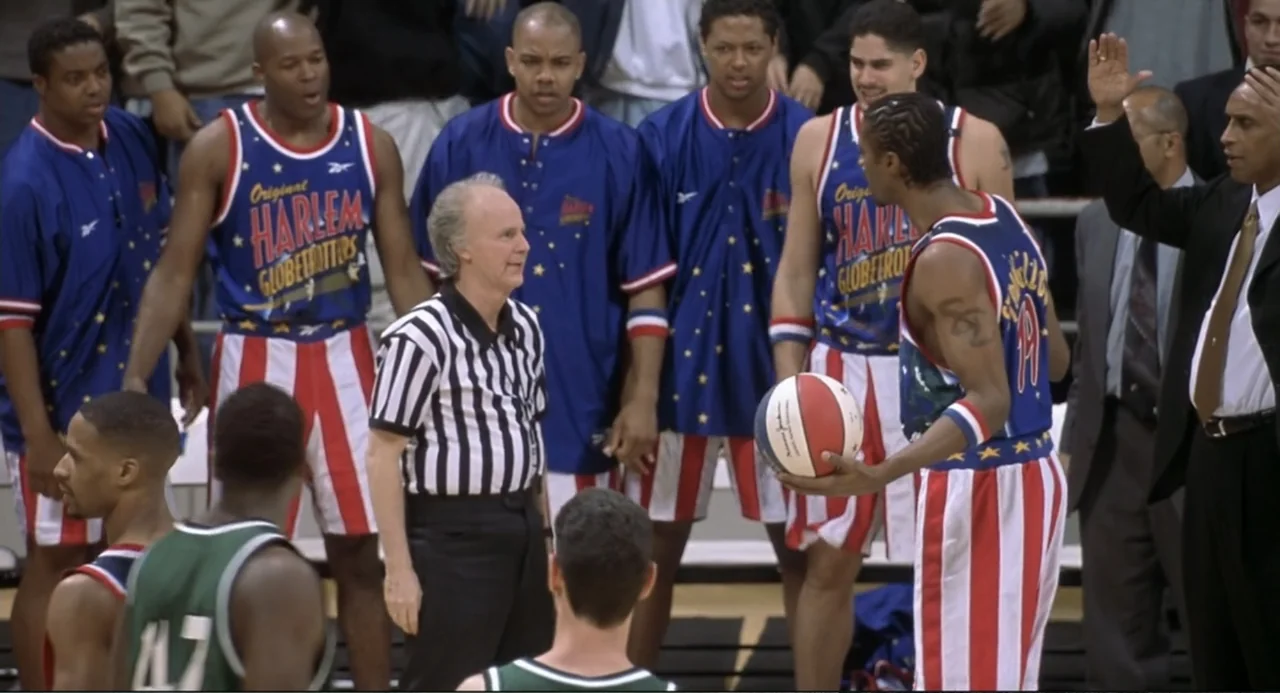 Little Nicky (2000) movie still: medium — An indoor basketball court features multiple athletes in team jerseys and a referee standing. The players…