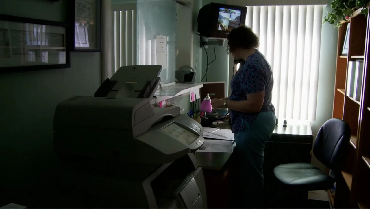 12th & Delaware (2010) movie still: medium — A cramped medical office features a large printer in the foreground and shelves nearby. A woman in…