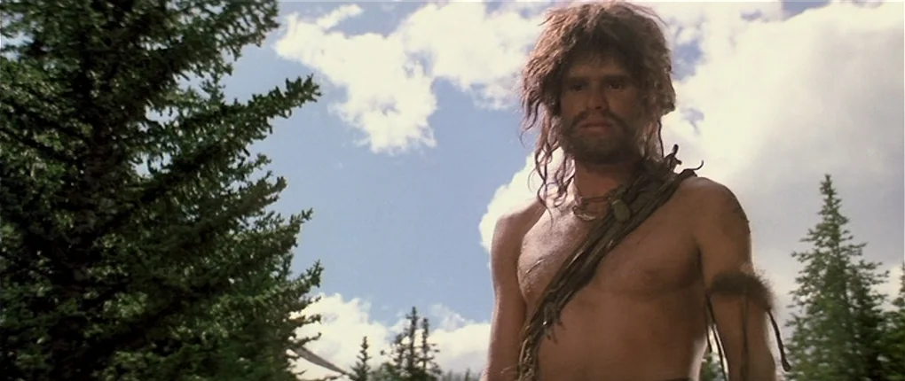 Clan of the Cave Bear (1986) movie still: medium — A dense coniferous forest with tall evergreen trees under a bright cloudy sky. A man with unkempt…