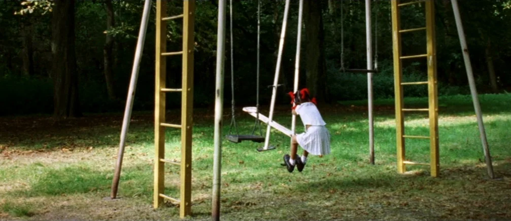 Innocence (2005) movie still: wide — A quiet outdoor area contains a metal teeter-totter structure set amidst mature trees. A young girl in a white…
