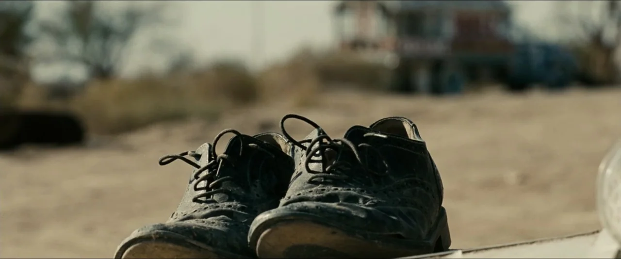 Into the Wild (2007) movie still: closeup — A parched desert landscape serves as the backdrop for a pair of discarded shoes. The leather dress shoes…