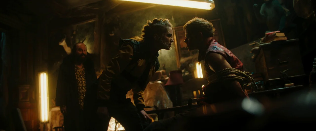 Boy Kills World (2023) movie still: medium — A cluttered room is filled with indistinct objects, antique furniture, and yellow-toned lighting. A…