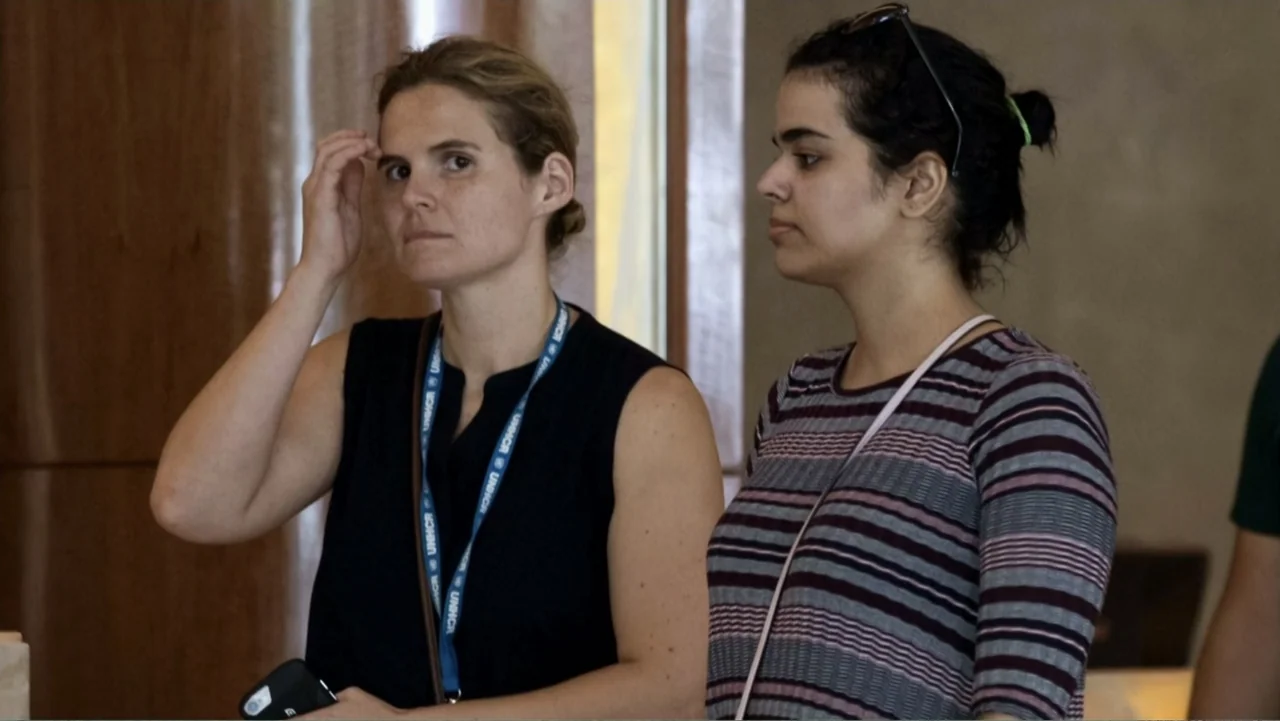 Escape From Saudi (2019) movie still: medium — An interior office space with plain walls and wooden accents. Two women stand next to each other, one…