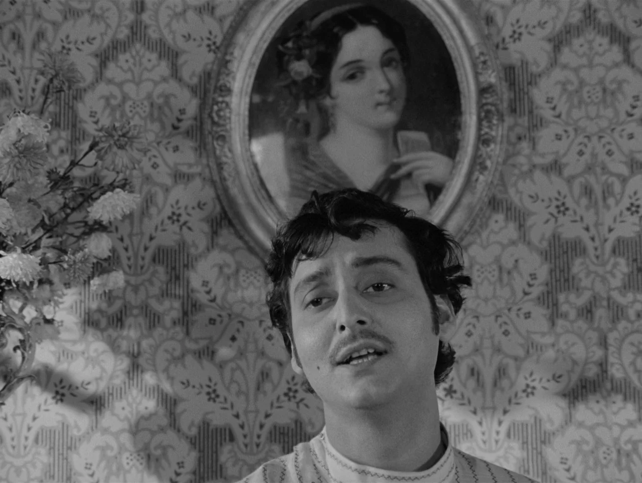 Charulata (1964) movie still: medium — An interior space features patterned floral wallpaper and a framed portrait of a woman. A man stands before…