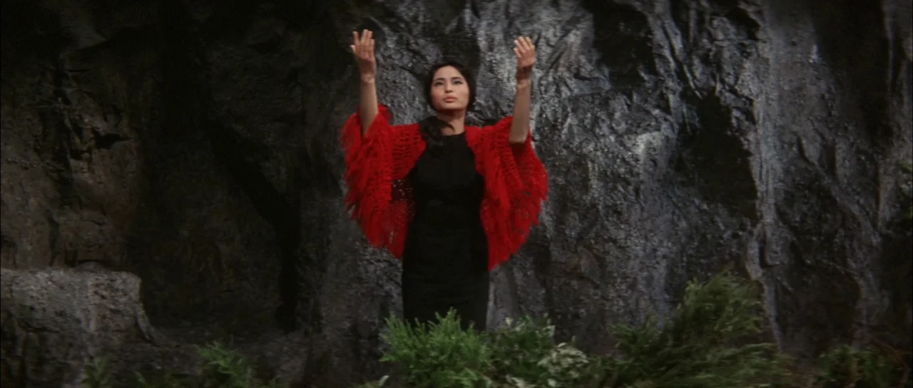 Ghidorah The Three Headed Monster (1964) movie still: medium — A woman stands in front of a dark, weathered rock wall outdoors. She is wearing a…