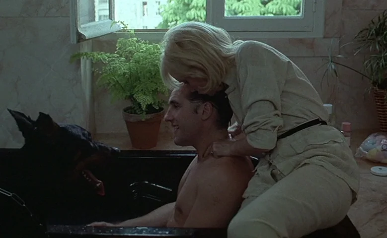 Maîtresse (1975) movie still: medium — A light-tiled space features a black tub set beneath a bright window with greenery. A woman in a light shirt…