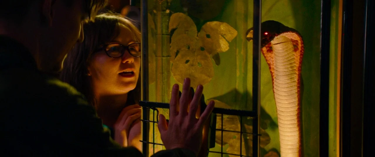 Dinner in America (2020) movie still: medium — An interior zoo exhibit display with green vegetation and a coiled snake inside glass. A woman and…