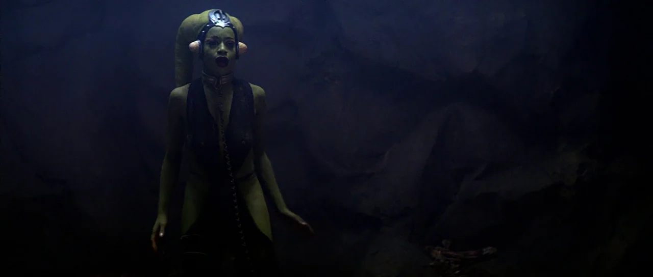 Return Of The Jedi (1983) movie still: medium — The scene is set inside a dark, shadowed cave with rocky, irregular walls. A green-skinned humanoid…