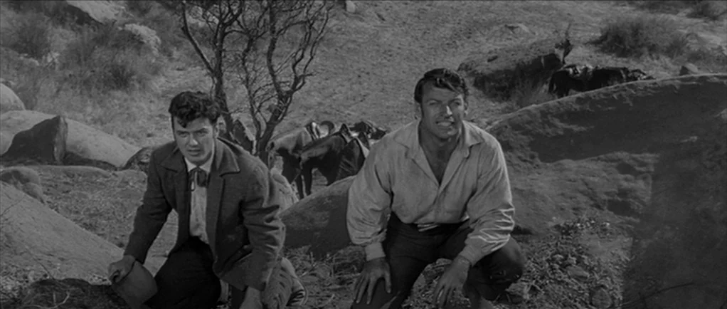 Love Me Tender movie still: medium — A desolate, sun-drenched rocky landscape with scrubby vegetation and horses in the background. Two men crouch…