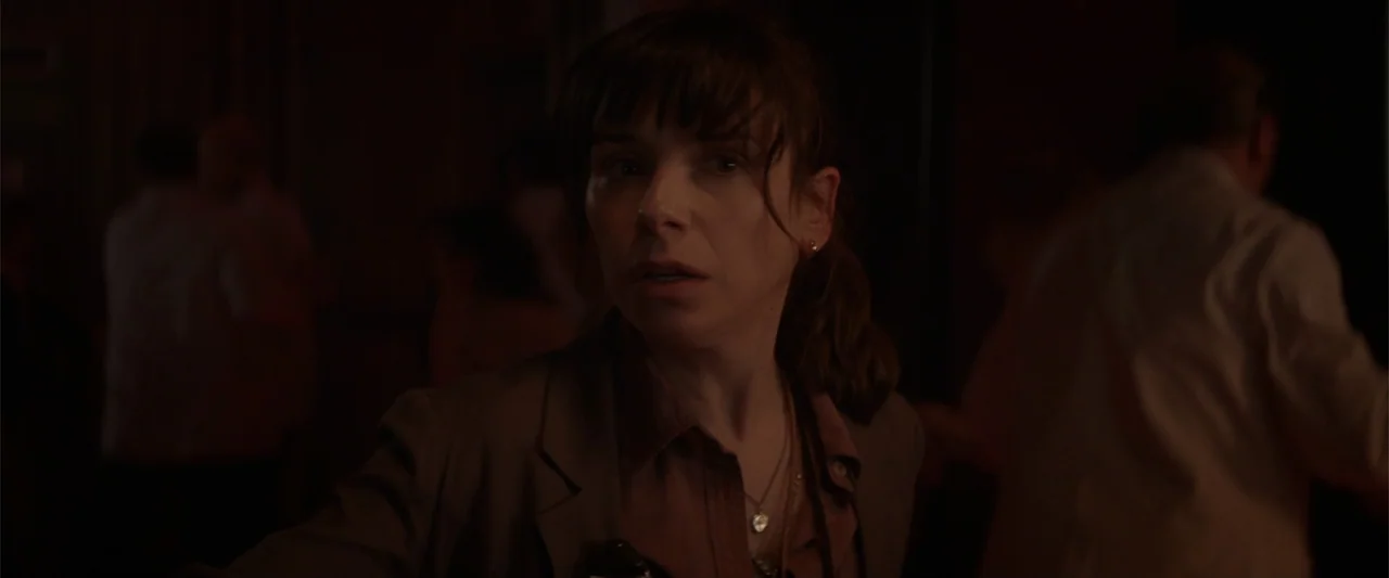 Godzilla (2014) movie still: closeup — A dimly lit, crowded interior space with blurred figures moving in the background. A woman in a dark blazer…