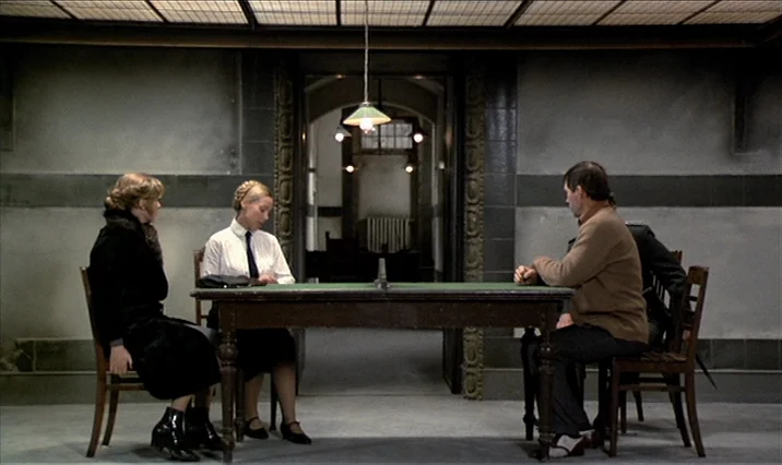 The Serpent’s Egg (1977) movie still: wide — An austere room features grey stone walls, tiled accents, and a central wooden table. Three people sit…