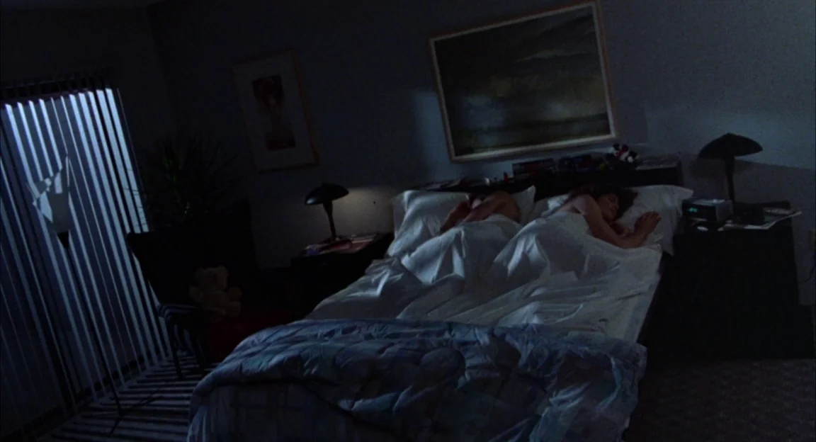 Creepshow 2 (1987) movie still: wide — A dark, dimly lit hotel room with two people sleeping in a large bed. Light from vertical blinds casts long…
