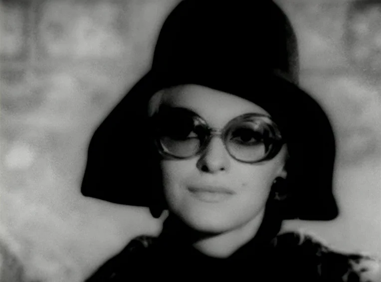 Cuadecuc, Vampir (1971) movie still: closeup — A close portrait of a woman in a dark hat and glasses. She faces forward against a blurry,…