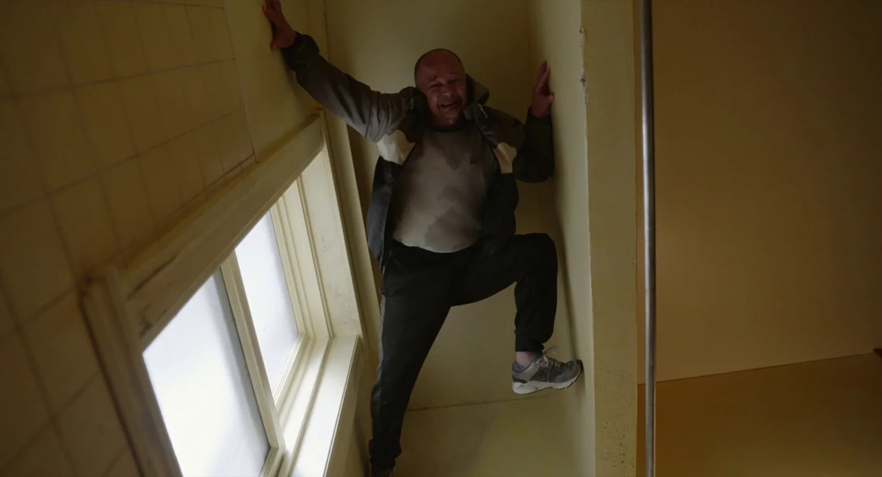 Beau Is Afraid (2023) movie still: fullbody — A narrow stairwell with yellow-painted walls and a large, bright window frame. A middle-aged man…