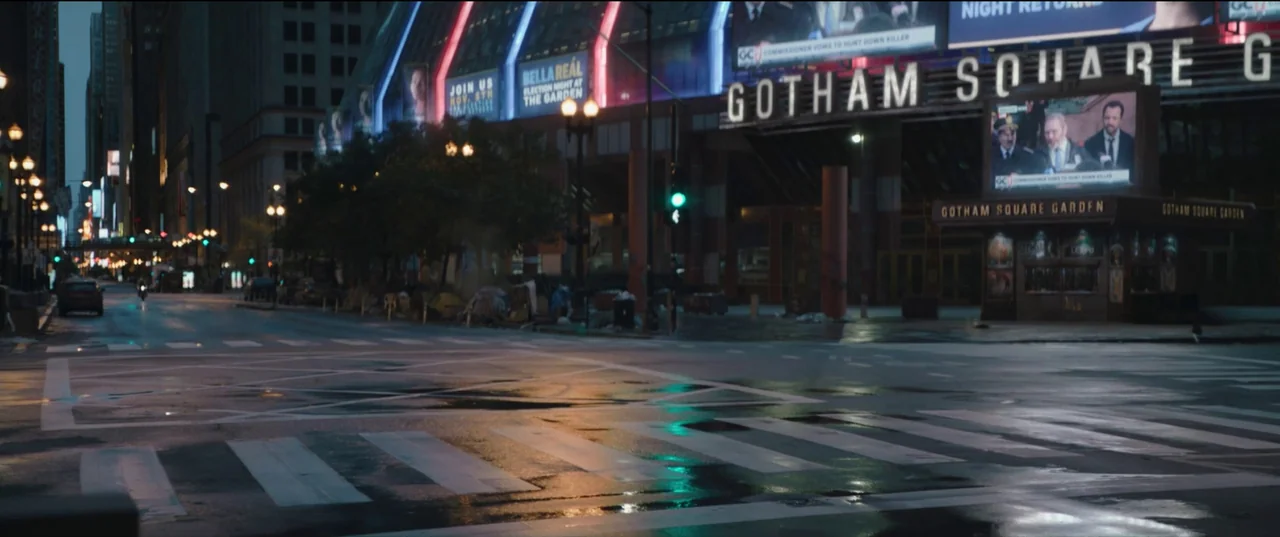 The Batman (2022) movie still: establishing — A wet city intersection features a large building complex with prominent lit signage. The street is…