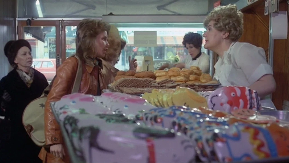 Sisters (1973) movie still: medium — A 1970s bakery features glass display cases filled with pastries and decorated cookies. A woman in a leather…
