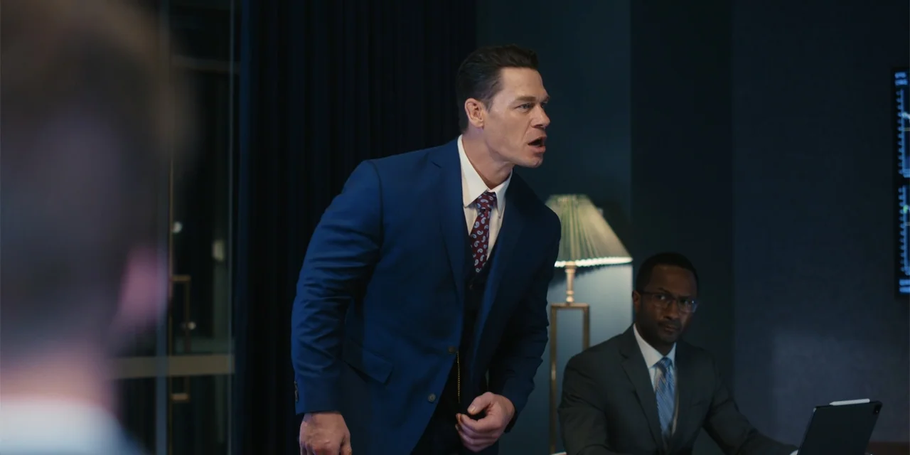 Ricky Stanicky (2024) movie still: medium — A dimly lit corporate environment features dark blue walls and a modern floor lamp. A standing man in a…