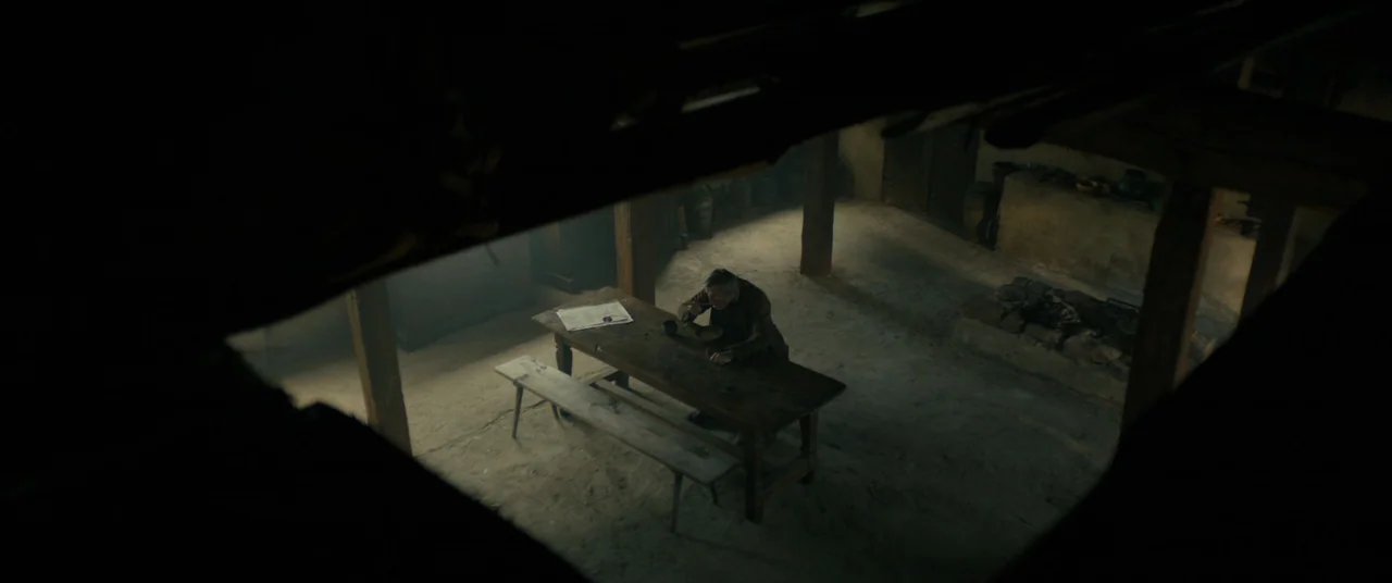 The Promised Land (2023) movie still: wide — A dark, rustic interior space features exposed wooden beams and an earthen floor surface. An elderly…