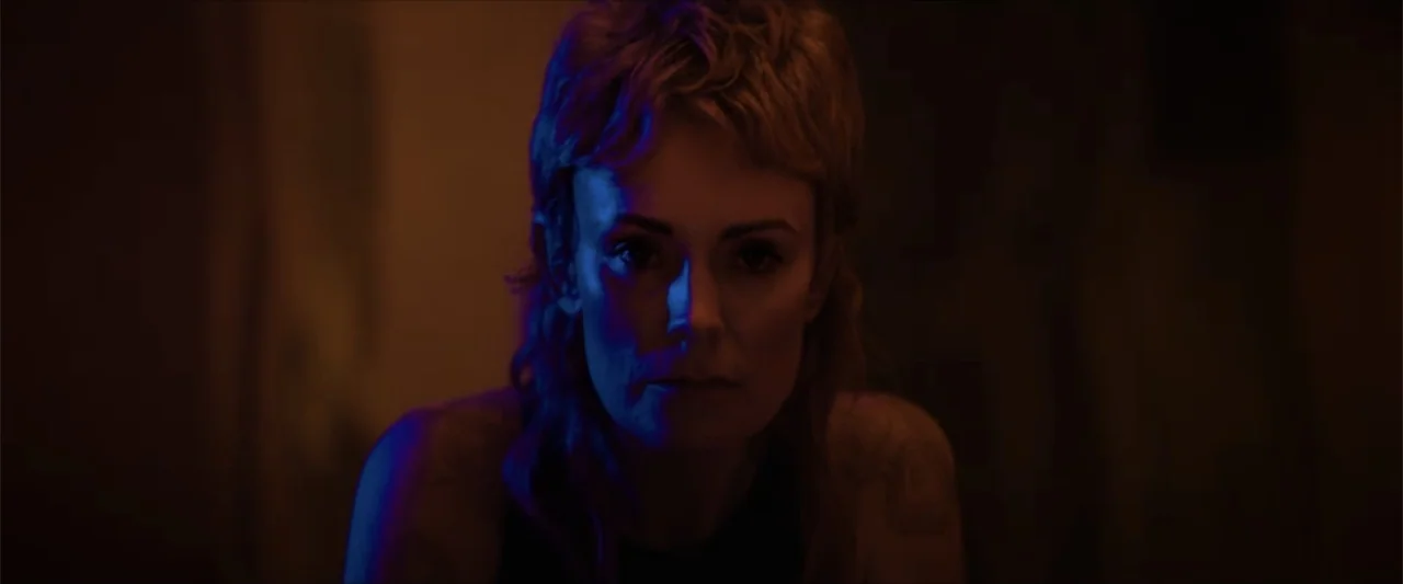 Ash (2025) movie still: medium — An interior space is dimly lit by contrasting colored light sources on the subject. A woman with short blonde hair…