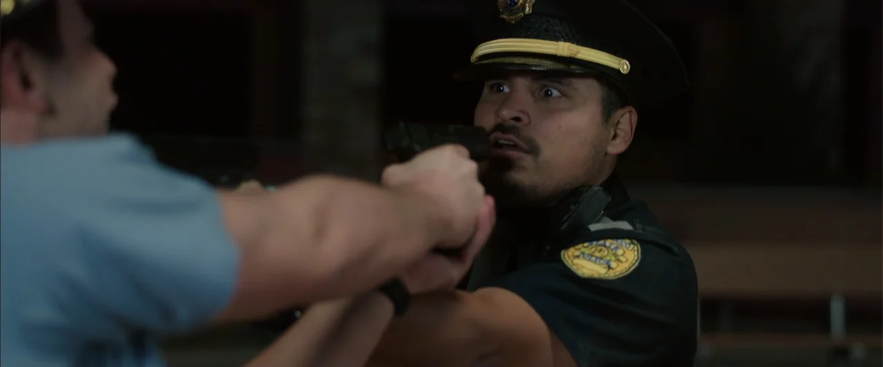 Vacation (2015) movie still: medium — A dark outdoor space at night with an out-of-focus background. A police officer has a handgun pointed directly…