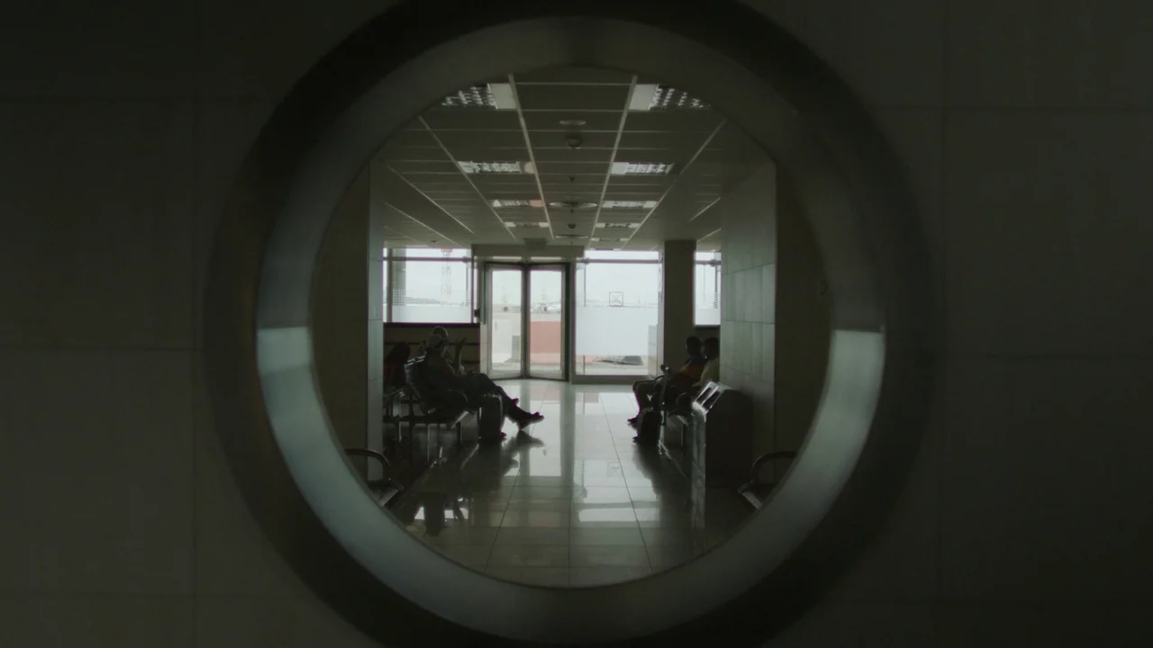 Infinity Pool (2023) movie still: wide — An airport waiting area viewed through a circular structural opening in a wall. Passengers are seated in…