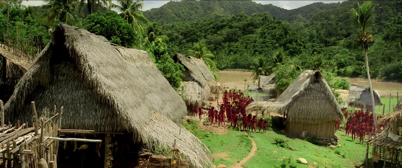 Green Inferno (2013) movie still: wide — A rural village of thatched-roof huts sits near a brown river in lush hills. A large group of people…
