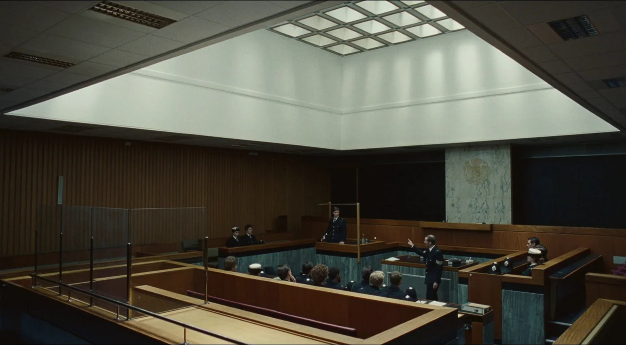 Red, White & Blue (2020) movie still: wide — A modern courtroom with wood-paneled walls and a large central skylight overhead. A police officer in…