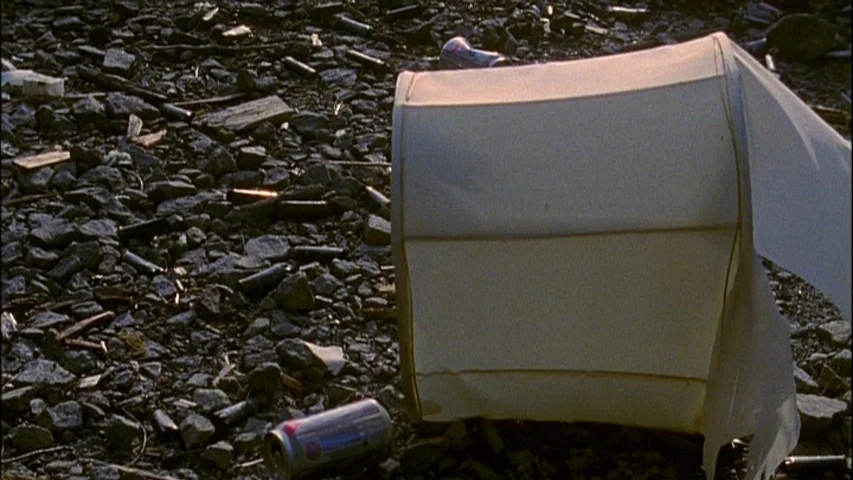 Old Joy (2006) movie still: medium — A discarded, damaged lampshade lies on a bed of dark, irregular stones and rocks. Scattered aluminum beverage…