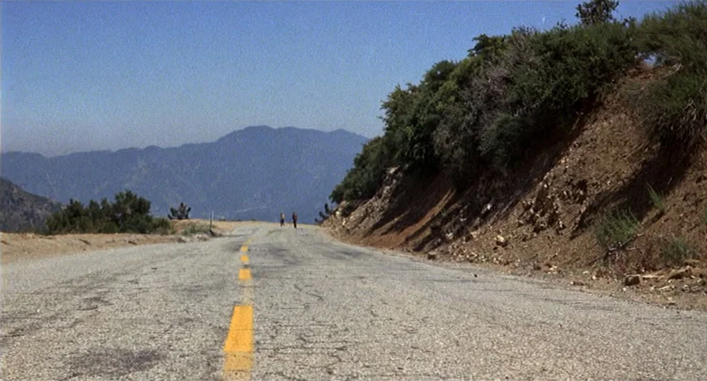 Family Plot (1976) movie still: wide — A cracked asphalt road winds through a dry, hilly mountainous landscape under clear skies. Two small figures…