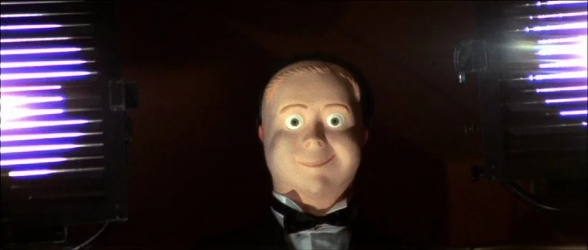 Four Flies On Grey Velvet (1971) movie still: closeup — A ventriloquist dummy stands against a dark background flanked by two bright light panels.…