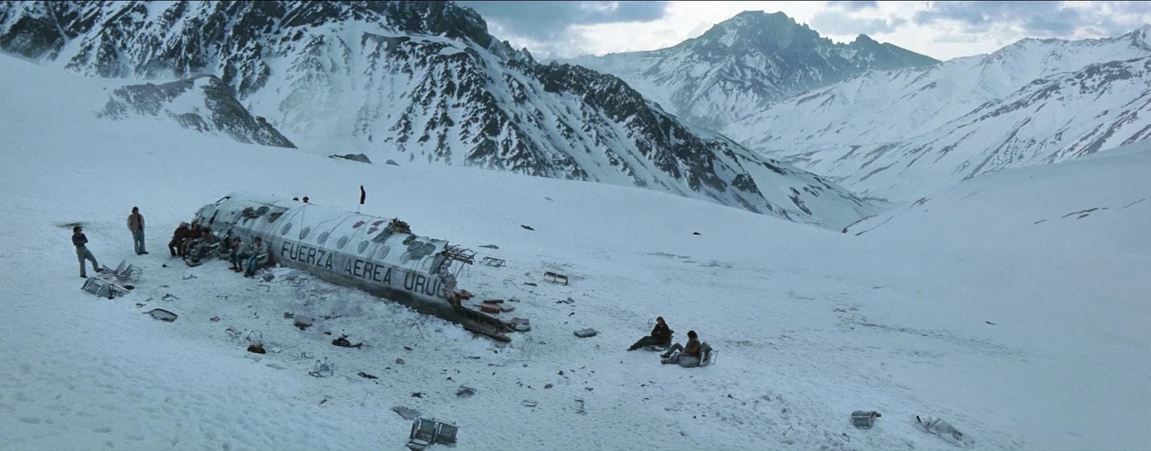 Society of the Snow (2023) movie still: establishing — A snow-covered mountain slope features the wreckage of a downed airplane fuselage. Several…