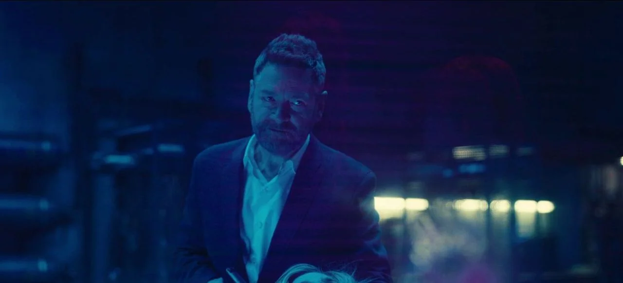 Tenet (2020) movie still: medium — An industrial space is bathed in intense blue light and deep shadows. A middle-aged man stands in the foreground…