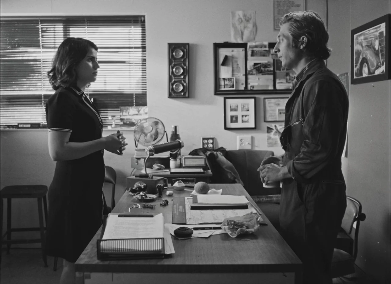 Fremont (2023) movie still: medium — An office space with a cluttered desk, a window with blinds, and framed art. A man and a woman stand opposite…