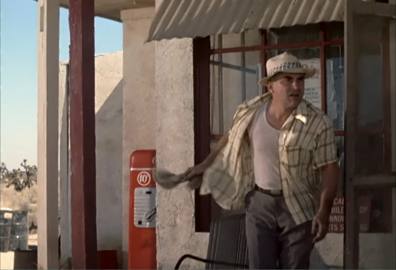 Black Cat Run (1998) movie still: medium — An arid, sun-bleached desert service station with a red fuel pump and stucco wall. A middle-aged man…