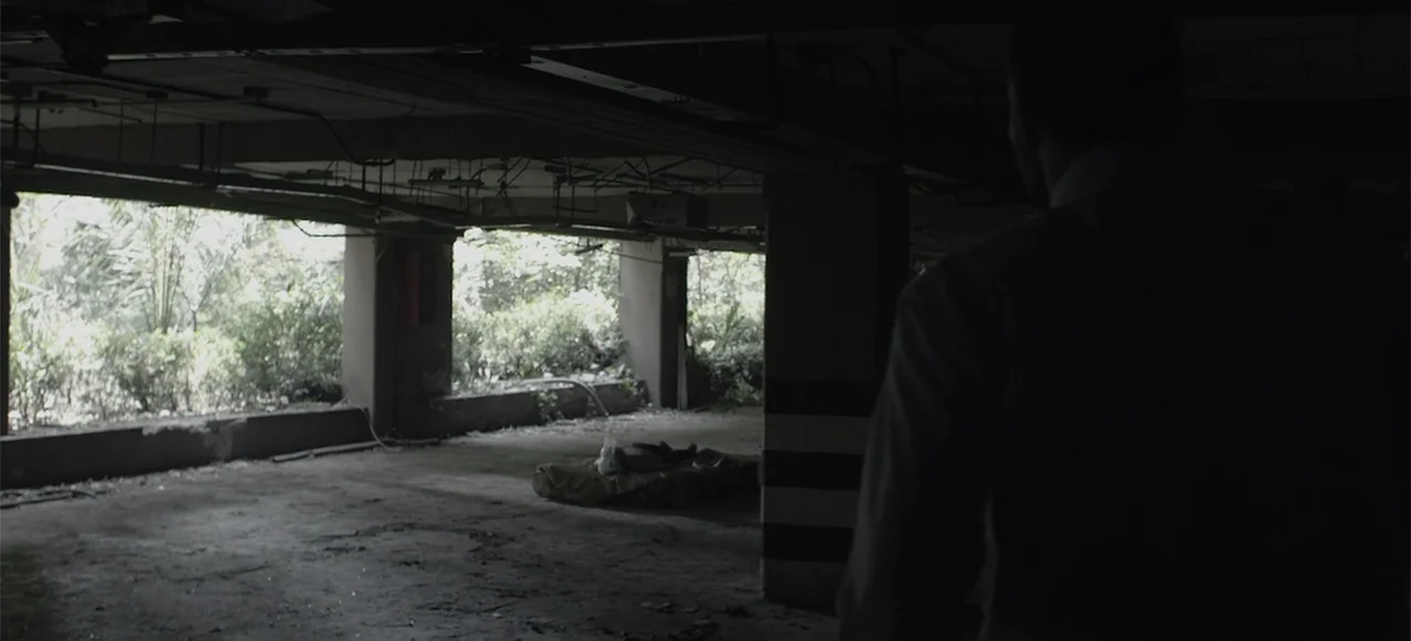 About the Deaths (2016) movie still: medium — A dimly lit concrete parking structure shows overgrown foliage through the open outer edge. A man…