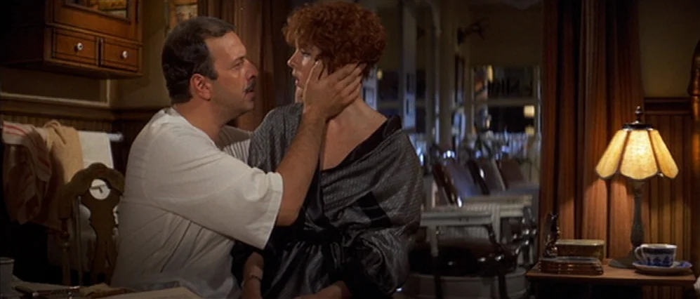 The Fortune (1975) movie still: medium — A dimly lit hotel room features dark wood paneling and vintage furniture. A man holds a woman's face while…