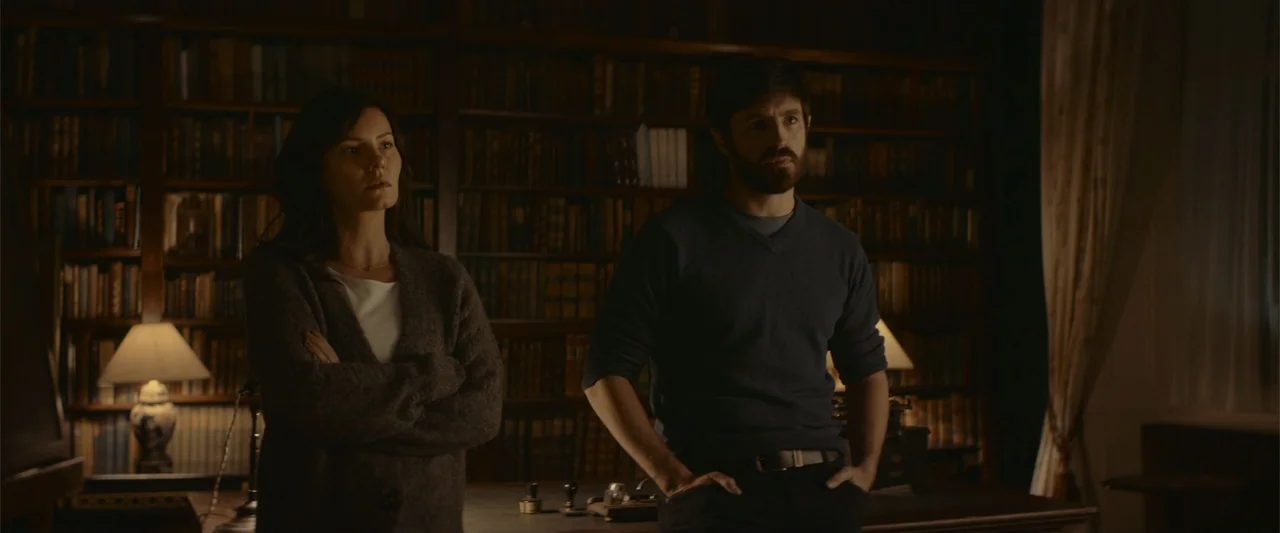 The Cellar (2022) movie still: medium — A dark library room features extensive floor-to-ceiling wooden bookshelves filled with aged volumes. A man…