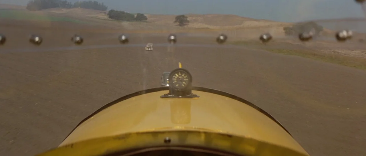The Rocketeer (1991) movie still: establishing — A dry, open landscape of tilled brown soil extends toward distant rolling hills. A yellow aircraft…