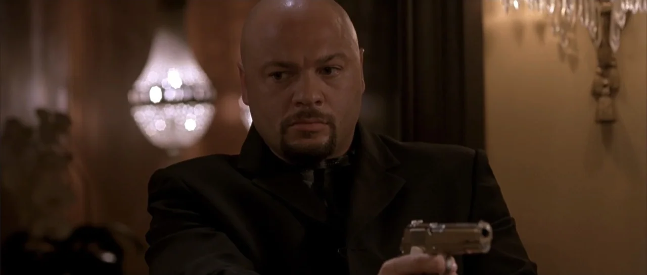 Shade (2003) movie still: medium — A dimly lit interior space features a chandelier blurred in the background. A man holds a silver handgun pointed…