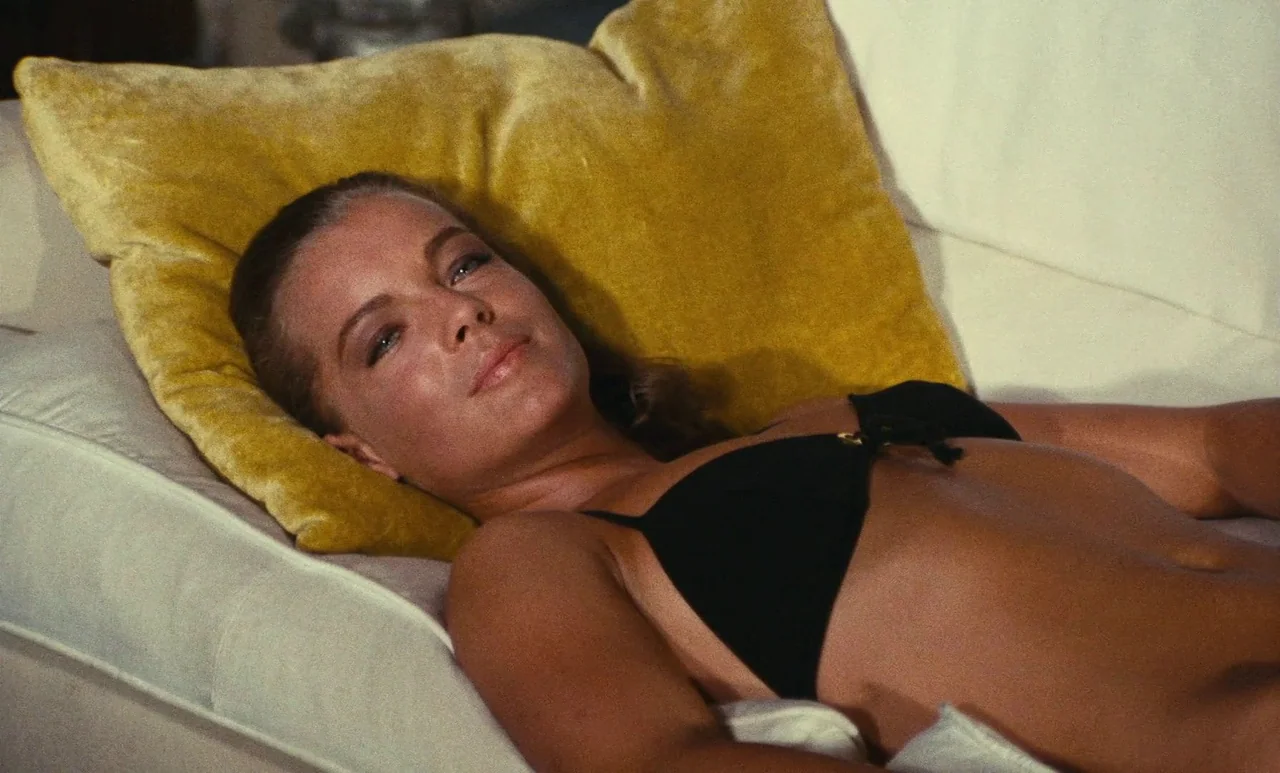 The Swimming Pool (1969) movie still: closeup — A woman rests on a plush white sofa with a vibrant yellow velvet pillow. She is lying on her back,…