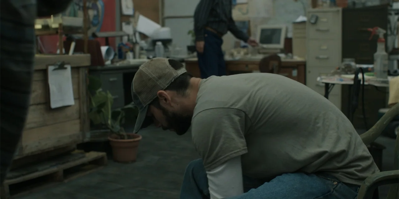 Caddo Lake (2024) movie still: medium — A cluttered workshop contains work tables, filing cabinets, a potted plant, and computer equipment. A man in…