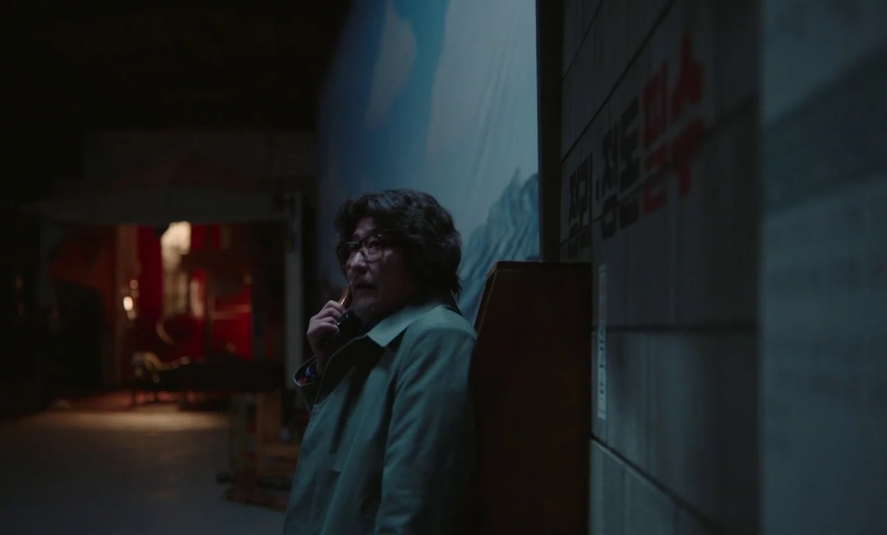 Cobweb (거미집) (2023) movie still: medium — A dimly lit indoor corridor features a mural wall with Korean text on the right. A man stands leaning…
