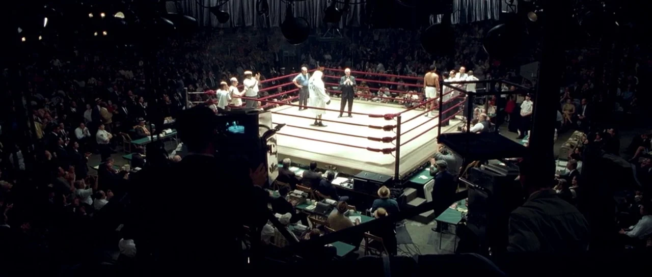 Ali (2001) movie still: wide — A high-angle view looks down upon a boxing ring at night in an arena. A referee stands between two boxers while the…
