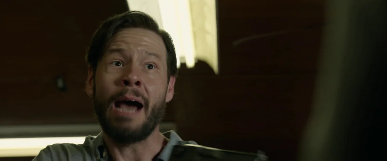 The Hunt (2020) movie still: closeup — A dark indoor space with brown wall paneling and rectangular ceiling light fixtures. A man in a blue shirt…