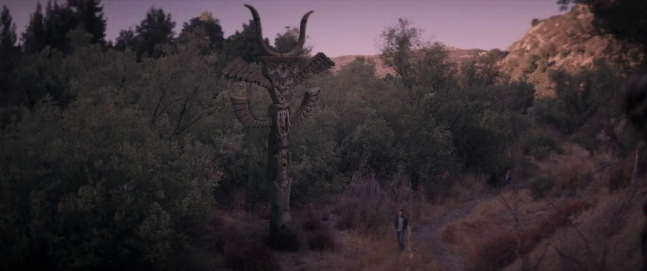 The Endless (2017) movie still: wide — A tall, elaborately carved wooden totem stands amidst dense, green shrubbery on a hillside. A man walks along…
