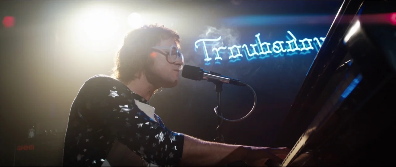 Rocketman (2019) movie still: medium — A dimly lit club stage features a neon Troubadour sign glowing in the background. A musician with…