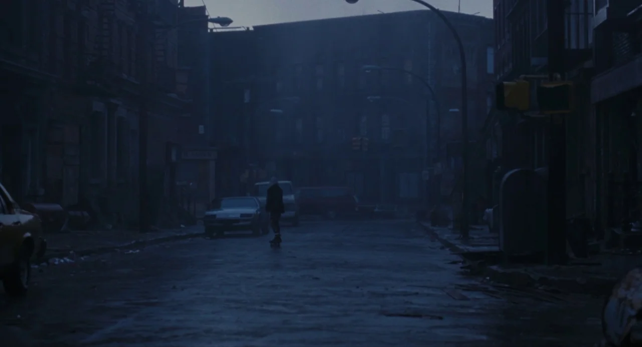The Crow (1994) movie still: wide — A dark, empty city street at night is lined with parked cars and buildings. A lone person on a skateboard rolls…