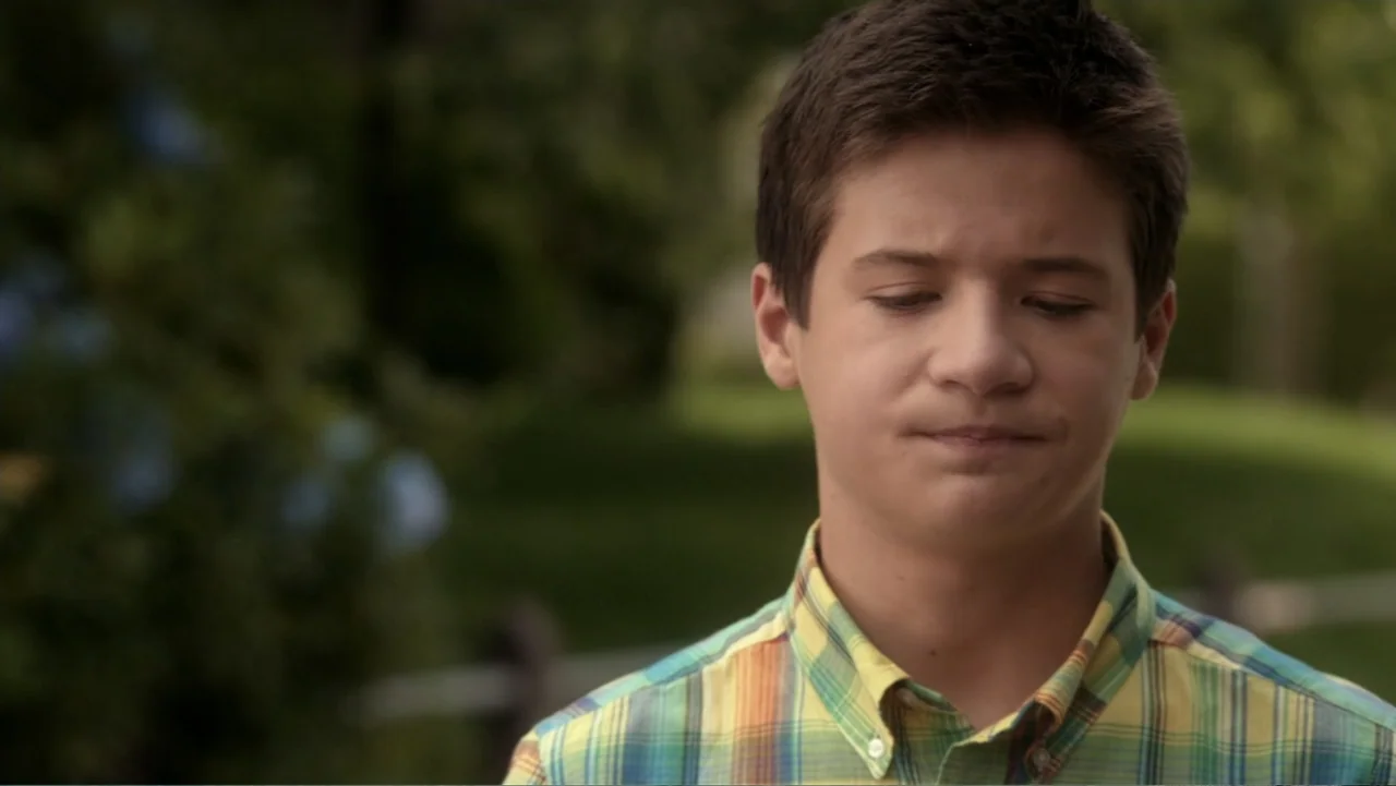 Rufus 2 (2017) movie still: medium — A green outdoor area features a blurred tree-filled background and white fence lines. A teenage boy in a plaid…