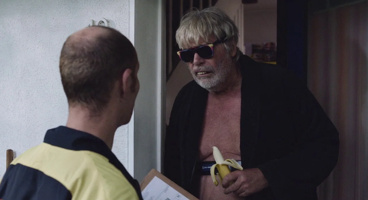 Toni Erdmann (2016) movie still: medium — A man in a bathrobe stands in a domestic doorway holding a peeled banana. He faces a delivery person who…